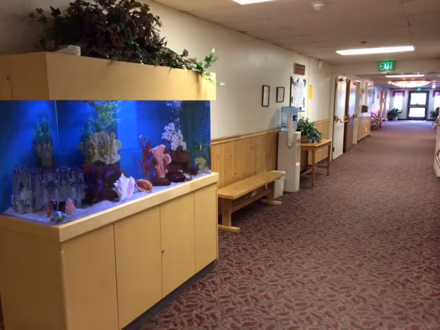 A long indoor hallway in a senior living facility with a large built-in aquarium on the left side filled with coral decorations and fish. The hallway has carpeted flooring with a patterned design, wooden paneling on the lower half of the walls, and a water cooler with a bench nearby. The hallway extends into the distance with doors and framed pictures on the walls, and an exit sign is visible at the far end.
