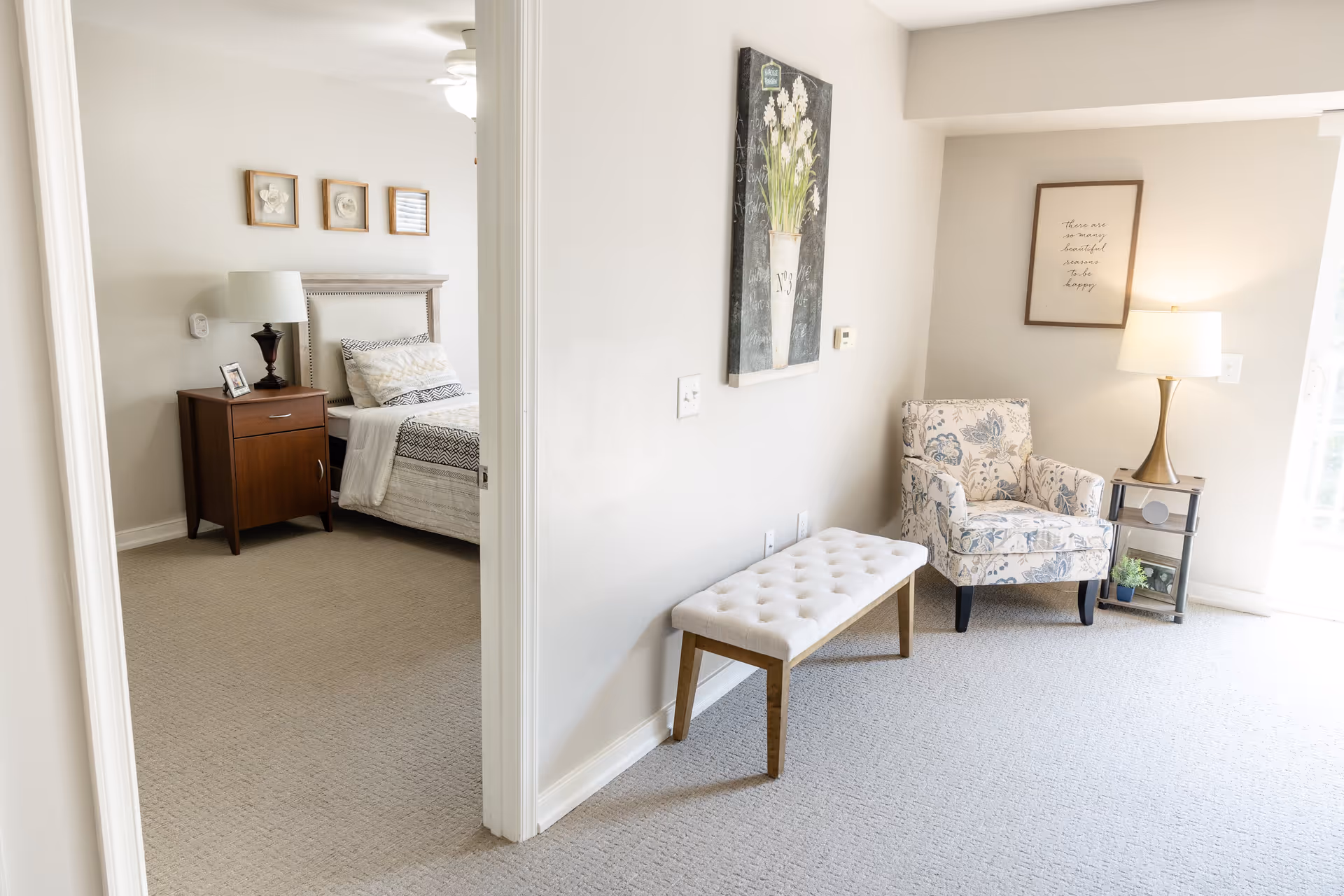 A bright and cozy interior space showing a small sitting area with a floral upholstered armchair, a side table with a lamp, and a cushioned bench against the wall. Adjacent to this area is a bedroom with a bed, nightstand, lamp, and framed pictures on the wall. The walls are light-colored and the floor is carpeted.