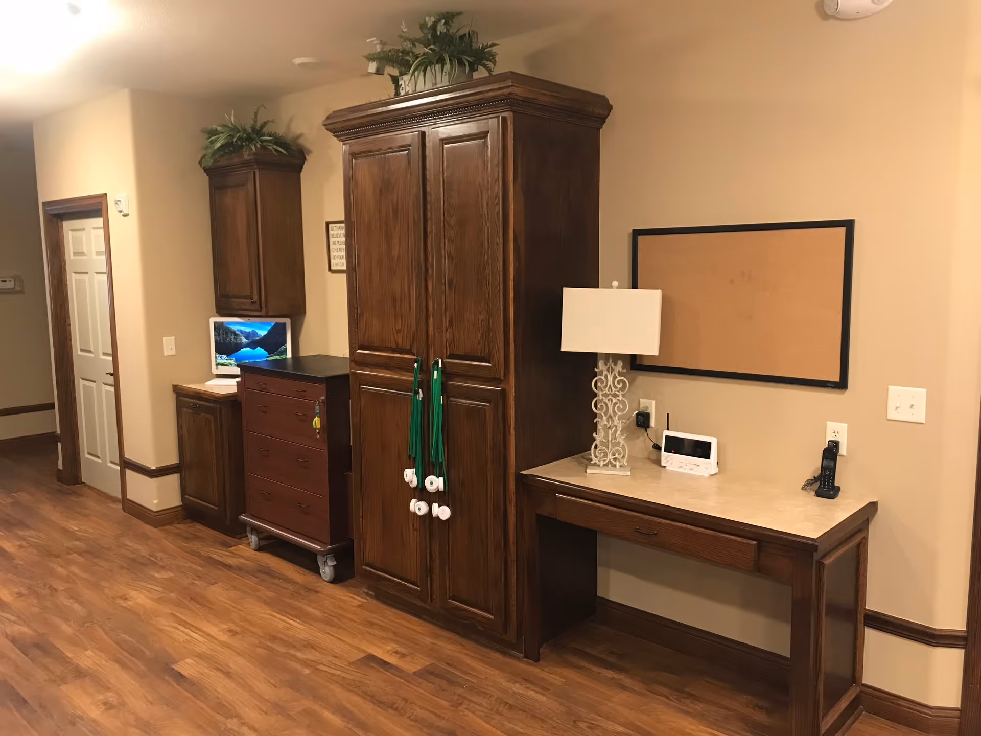 Interior hallway area with wooden cabinets and a desk. The cabinets are dark wood with plants on top. A small TV is on a cabinet, and a lamp, phone, and electronic device are on the desk. There is a corkboard on the wall above the desk. The floor is wood, and the walls are beige.