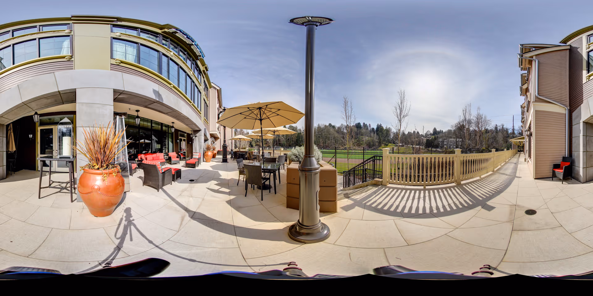 Outdoor patio area at Aegis Living Queen Anne Rodgers Park featuring seating arrangements with red cushions, tables with umbrellas, large potted plants, and a view of a green lawn and trees in the background under a clear sky.