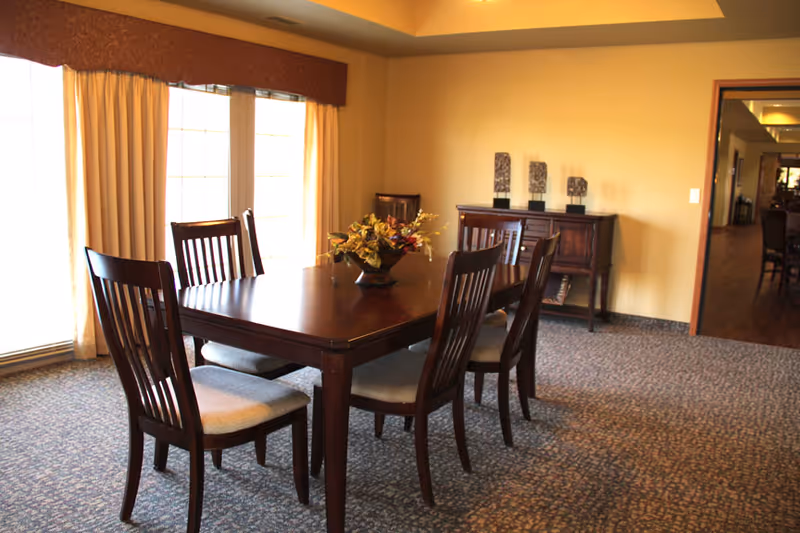 A formal dining room with a wooden table and six chairs, a floral centerpiece, sideboard and large curtained windows.