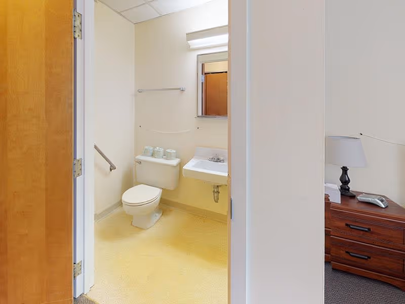 View into a small bathroom with a toilet, a wall-mounted sink, a mirror above the sink, and a towel rack on the wall. Part of a wooden door and a bedside table with a lamp and a phone are visible outside the bathroom.