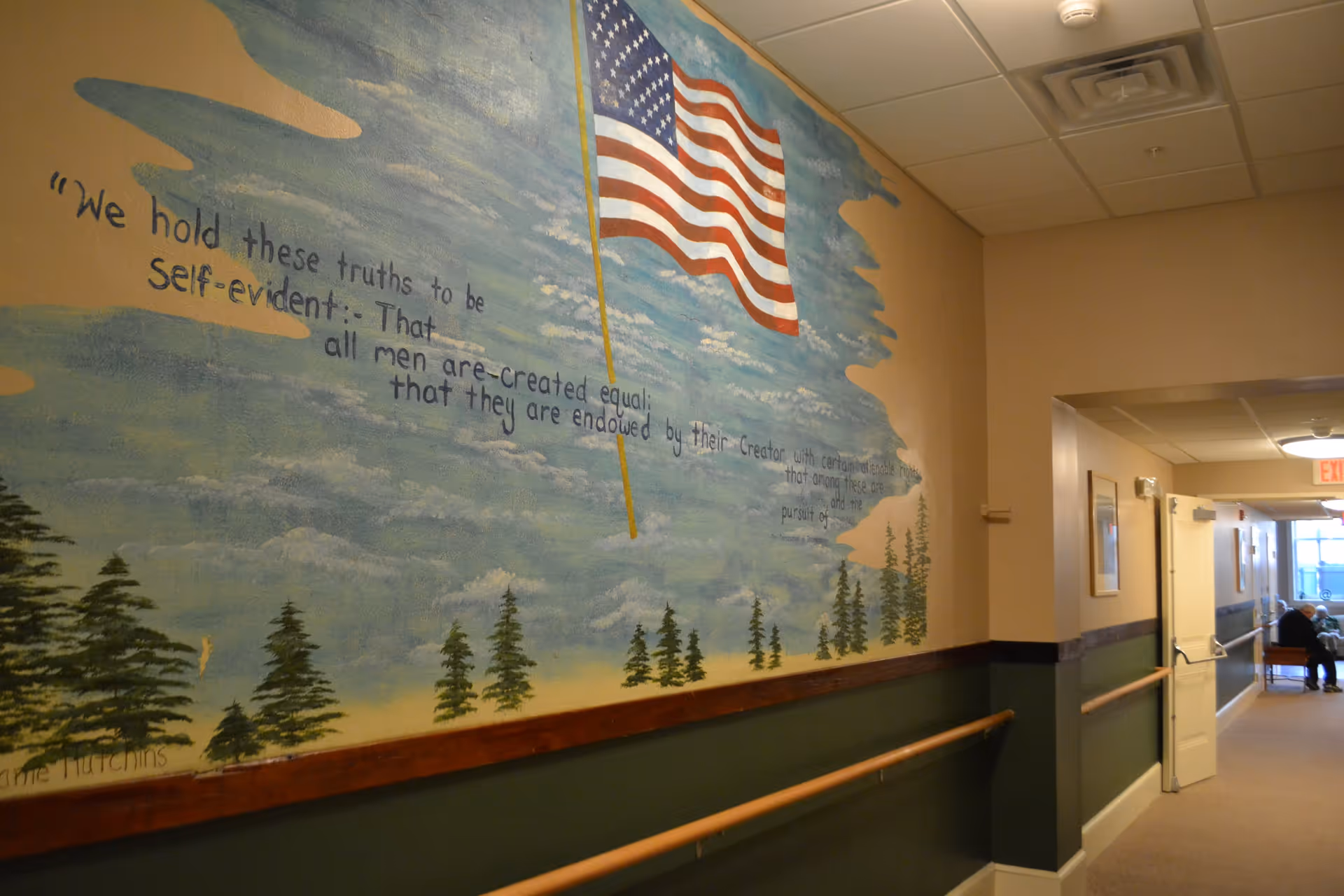 A hallway in a senior living facility with a mural on the wall depicting an American flag waving in the sky above a forest of pine trees. The mural includes a quote from the Declaration of Independence: 'We hold these truths to be self-evident: That all men are created equal; that they are endowed by their Creator with certain unalienable Rights...'. The hallway has handrails on both sides and leads to a seating area where a person is sitting near a window.