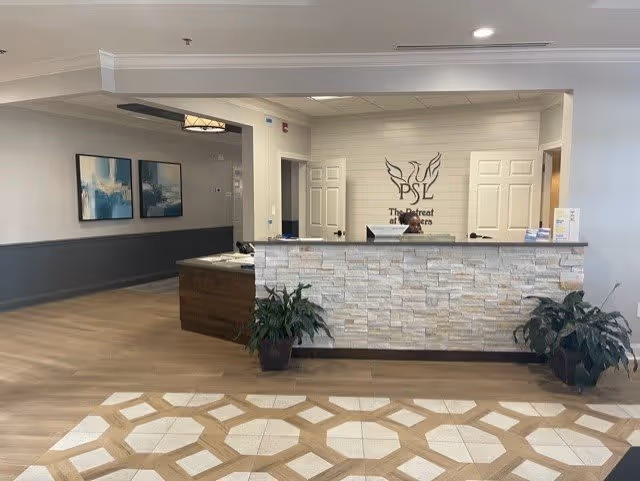 Reception area of The Retreat at Conyers featuring a stone front desk with a person seated behind it. The wall behind the desk displays the facility's logo and name. There are two potted plants on either side of the desk, wood and tile flooring, and two abstract paintings on the left wall.