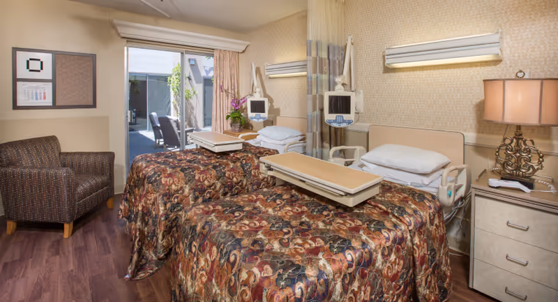 A senior living facility bedroom with two hospital-style beds covered in patterned bedspreads. Each bed has a pillow and an adjustable overbed table. There are medical monitors attached to the beds. A beige armchair is placed near the wall on the left side. The room has a sliding glass door leading to an outdoor patio area with chairs. A nightstand with a lamp and a phone is on the right side of the beds. The walls are decorated with a framed picture and a large mirror.