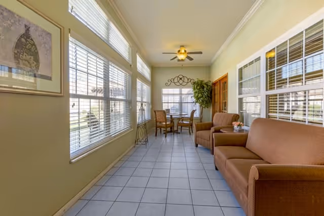 A bright sunroom with large windows on both sides, featuring a brown sofa and armchair on the right, a small table with a flower vase, and a round dining table with four chairs at the far end. The room has tiled floors, light yellow walls, a ceiling fan with light, and decorative wall art and a potted plant near the dining area.