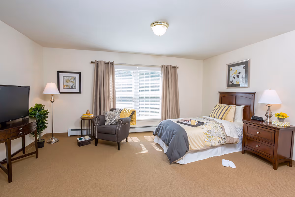 A well-lit bedroom with a single bed featuring a patterned comforter and pillows. Next to the bed is a wooden nightstand with a lamp, a clock, and a small vase with yellow flowers. A pair of white slippers is placed on the carpeted floor beside the bed. Across from the bed is a dark gray armchair with decorative pillows and a yellow throw blanket, positioned near a window with beige curtains and white blinds. A small round side table with books and a decorative item is next to the armchair. A flat-screen TV sits on a wooden console table, and a floor lamp and a potted plant are placed nearby. The walls are light-colored with framed artwork.