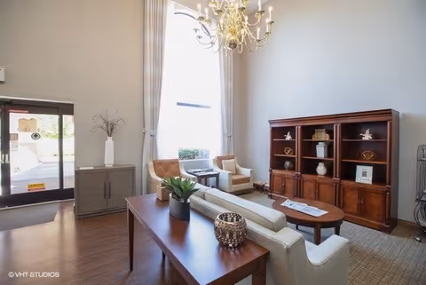 A bright and spacious living room area with high ceilings and a large arched window letting in natural light. The room features a white sofa, two beige armchairs, a wooden coffee table, a wooden console table with decorative items, and a large wooden bookshelf with various decorations. A chandelier hangs from the ceiling, and there is a glass door leading outside.