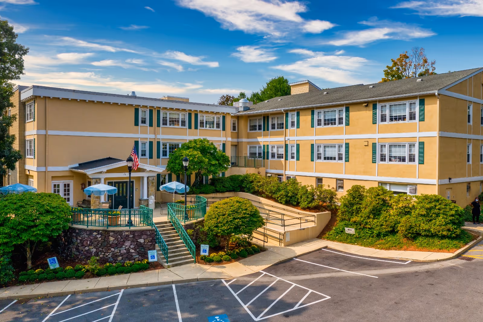 Exterior view of a three-story beige building with green shutters and white trim, surrounded by green bushes and trees. There is a small patio area with tables and blue umbrellas, an American flag near the entrance, and a parking lot with marked spaces in front of the building under a partly cloudy sky.