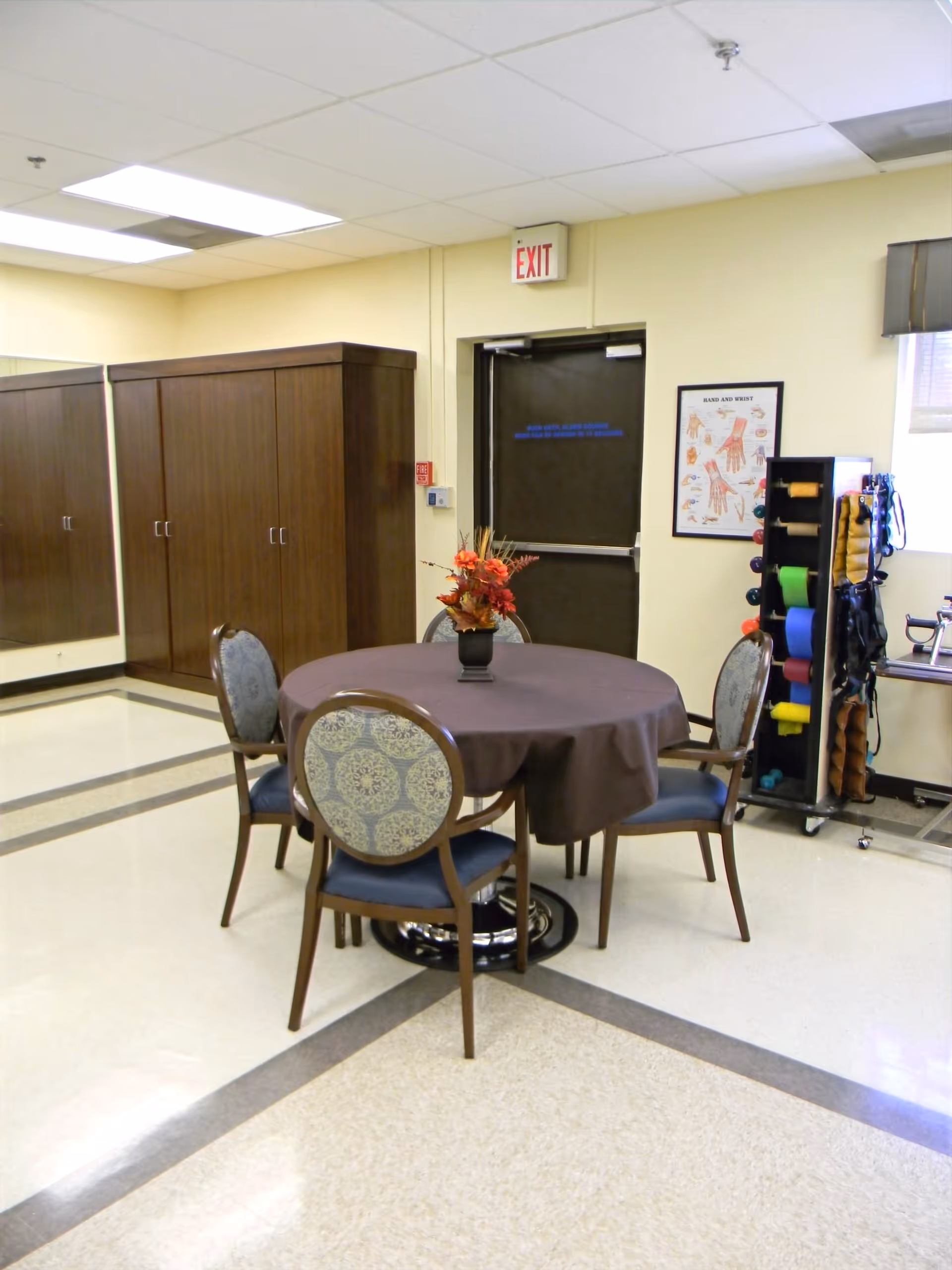 A room with a round table covered with a brown tablecloth and four upholstered chairs around it. There is a floral centerpiece on the table. In the background, there is a dark brown door with an exit sign above it, a wooden cabinet, a rack with colorful exercise bands, and a poster showing hand and wrist anatomy on the wall.