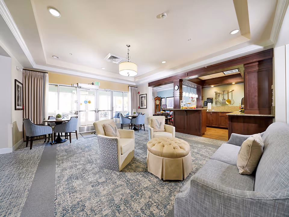 Bright communal lounge with sofas, armchairs, tables, and a small serving bar near glass entry doors.