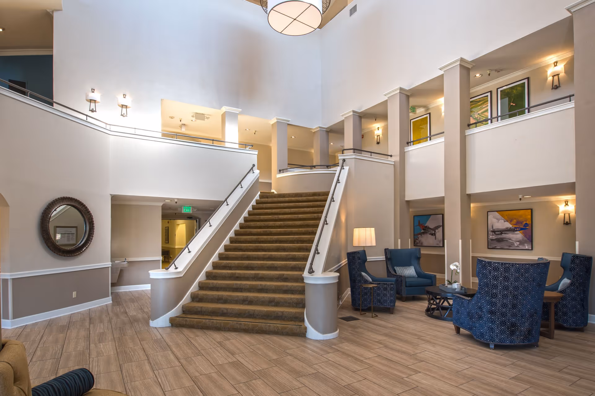 Spacious interior of a senior living facility featuring a wide carpeted staircase with handrails leading to an upper floor. The area includes a seating arrangement with blue upholstered chairs and a small table with a lamp and flower vase. The walls are decorated with framed artwork and a round mirror, and the space is well-lit with wall sconces and a large ceiling light fixture.