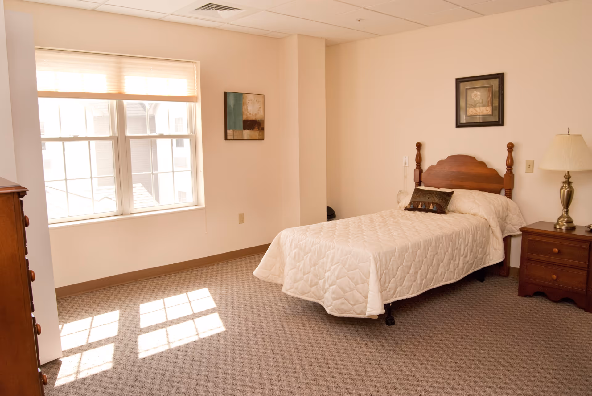 A simple, well-lit bedroom with a single bed covered in a white quilted bedspread. The room has beige walls and carpeted floor. There is a wooden nightstand with a lamp on the right side of the bed and a wooden dresser on the left side near a large window with blinds partially drawn. Two framed pictures hang on the walls.