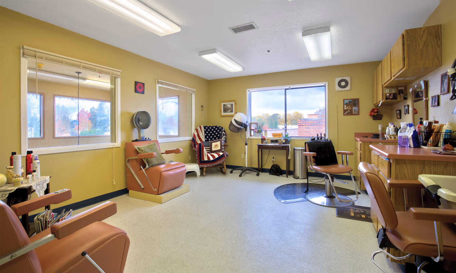 Interior of a hair salon room in a senior living facility with several salon chairs, a hair dryer, a large window showing an outdoor view, and shelves with hair care products and supplies.