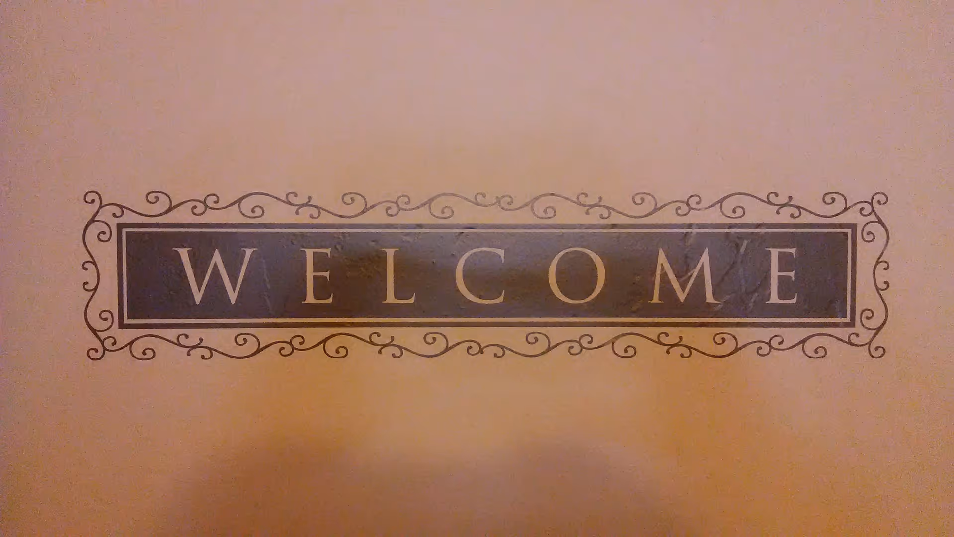 A decorative wall sign with the word 'WELCOME' in capital letters, framed by an ornate border with swirls and curls on a beige wall.