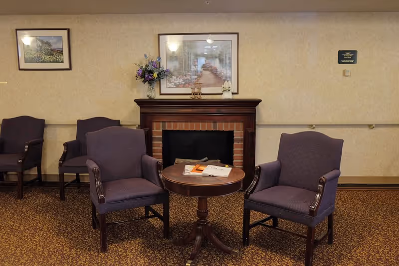 A cozy sitting area with two purple upholstered armchairs facing a small round wooden table with magazines on it. Behind the table is a brick fireplace with a wooden mantle, decorated with a vase of flowers and a small figurine. Two framed pictures hang on the beige wallpapered wall above the fireplace and to the left. The floor is covered with a patterned carpet.