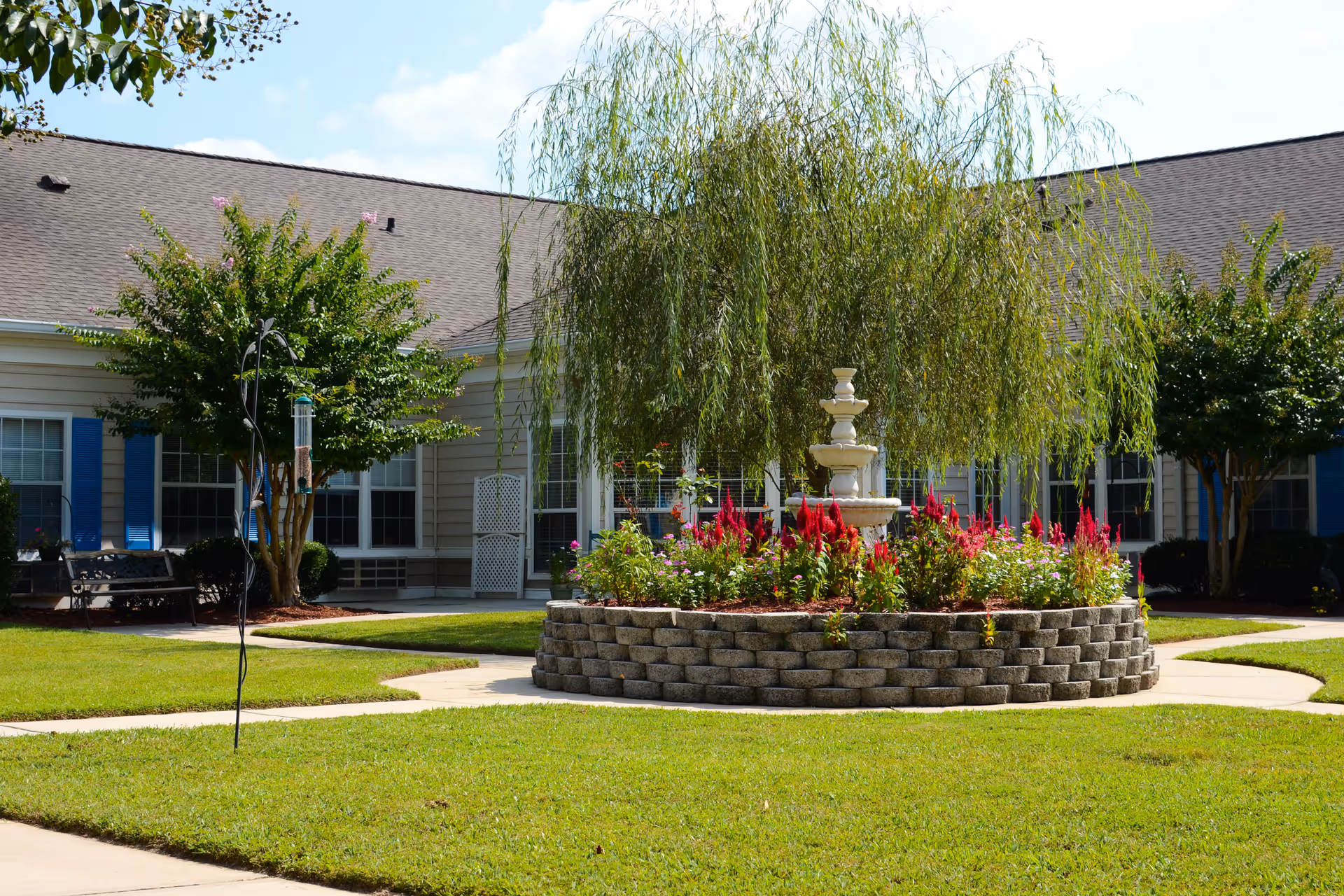 Outdoor courtyard area at The Legacy of Orangeburg featuring a circular raised flower bed with a multi-tiered white fountain in the center, surrounded by colorful flowers and a weeping willow tree. The courtyard is bordered by a building with beige siding, blue shutters, and multiple windows. There are paved walkways and green grass around the flower bed.