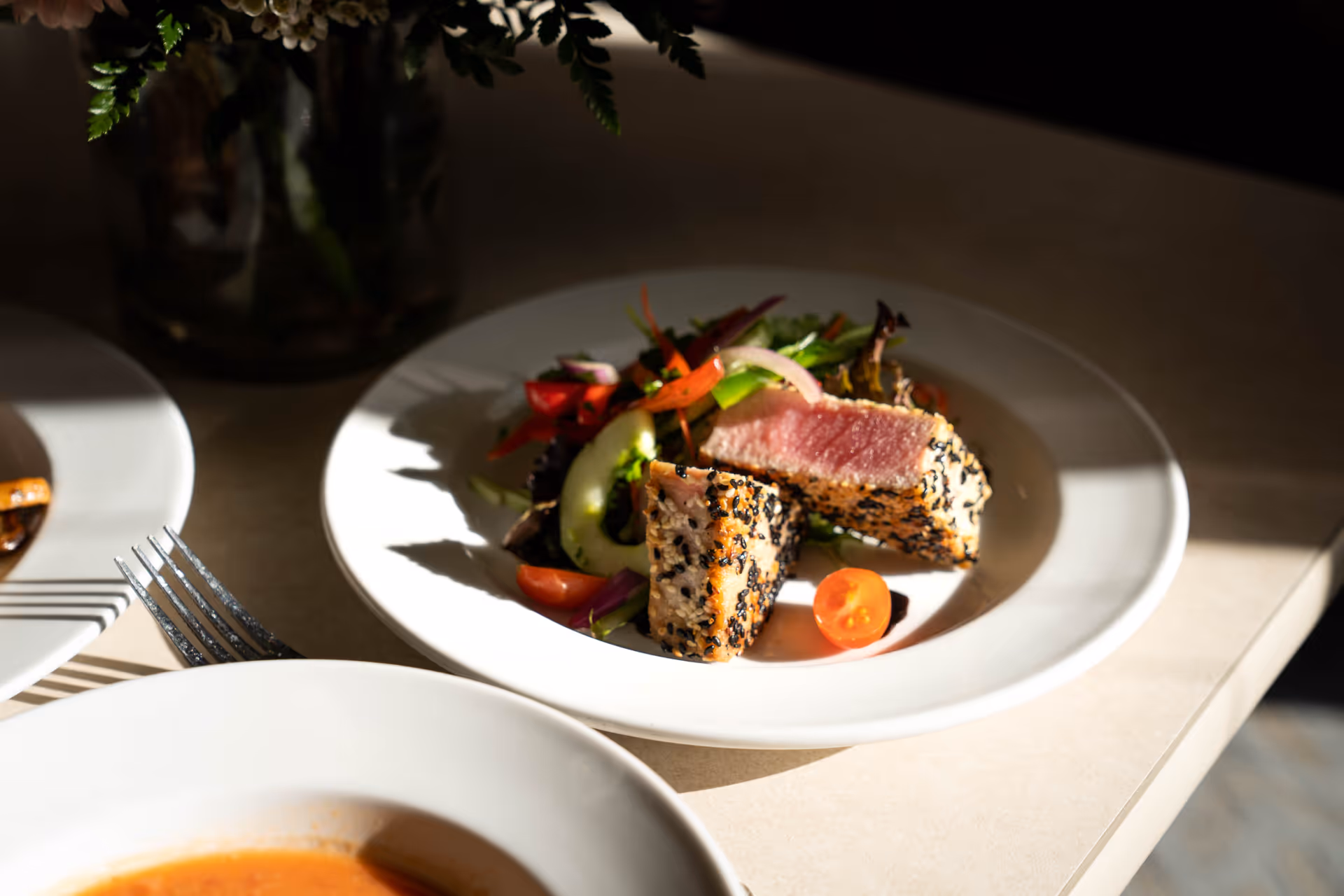 A white plate with a serving of seared sesame-crusted tuna, sliced to show the pink interior, accompanied by a fresh salad with cherry tomatoes, cucumber, and mixed greens on a light-colored table.