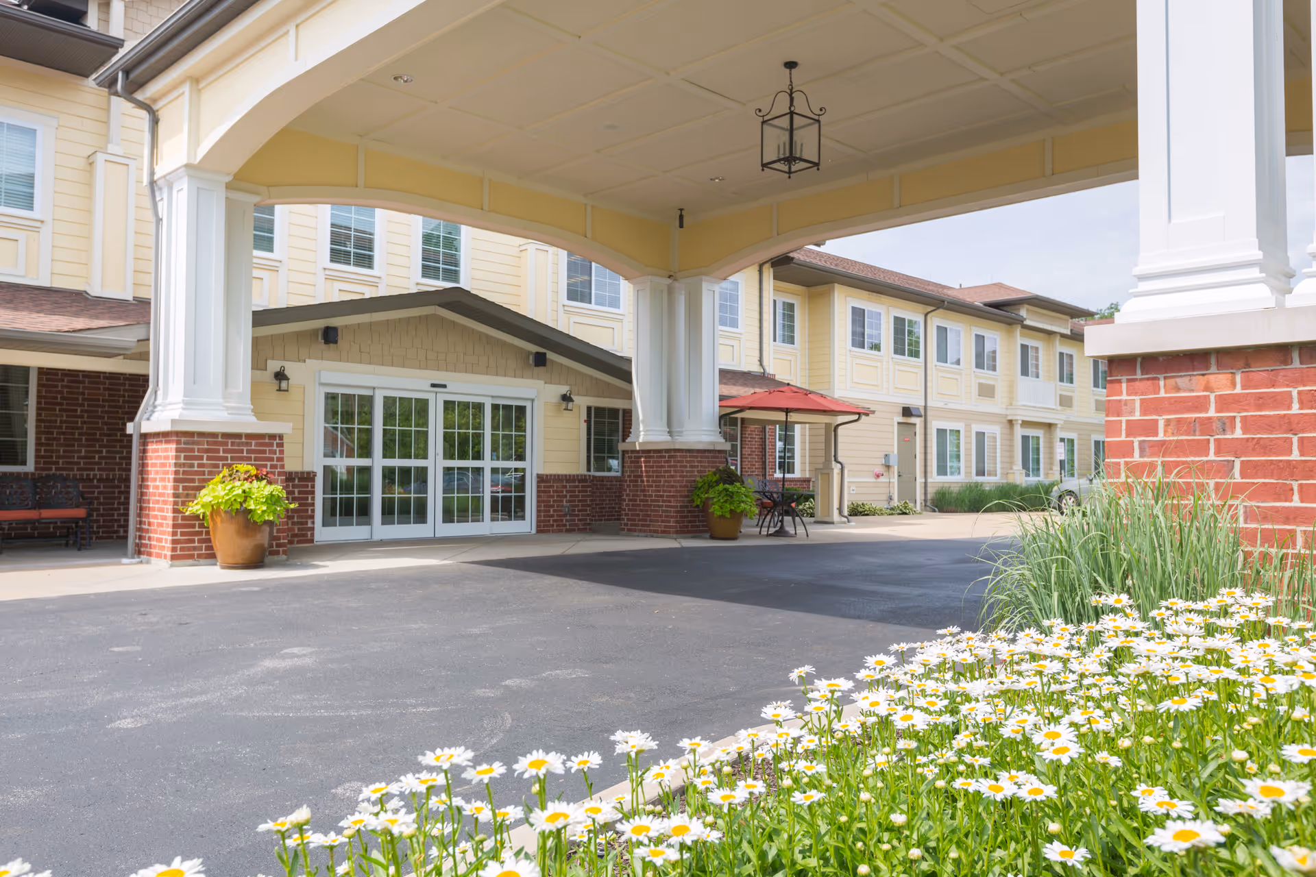 Entrance of Southview Assisted Living & Memory Care facility showing a covered drop-off area with white columns and brick accents. There are potted plants near the automatic sliding glass doors, a small table with a red umbrella, and a flower bed with white daisies in the foreground.