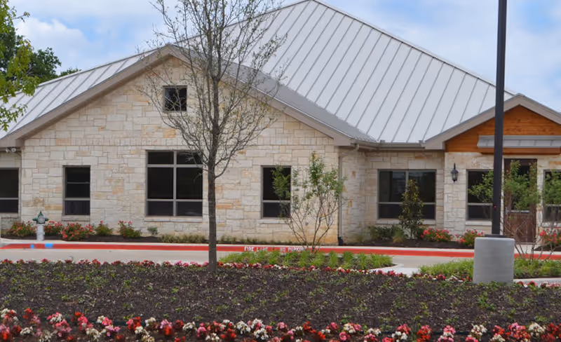 Front exterior of a single-story stone-faced assisted living building with a metal roof, landscaped flower beds, and small trees.