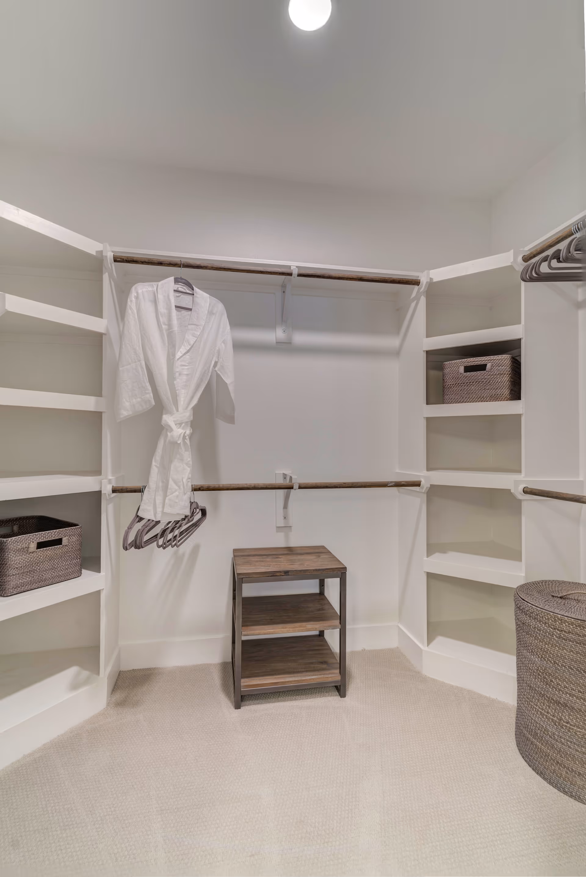 A walk-in closet with white walls and carpeted floor featuring built-in white shelves and hanging rods. There is a white robe hanging on a hanger, several empty hangers, a small wooden shelving unit in the center, and woven storage baskets on the shelves and floor.