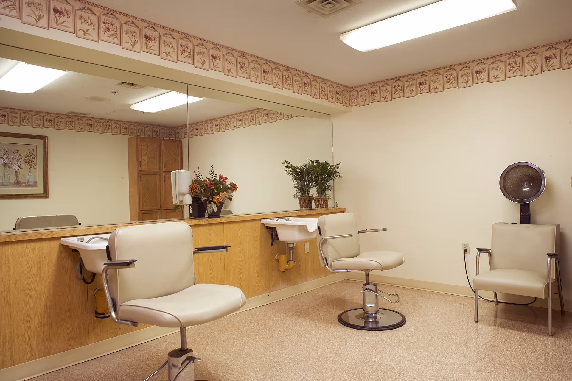 Interior of a salon area with two white salon chairs in front of sinks, a large mirror on the wall, floral wallpaper border near the ceiling, a hair dryer chair, and decorative plants and flowers on the counter.