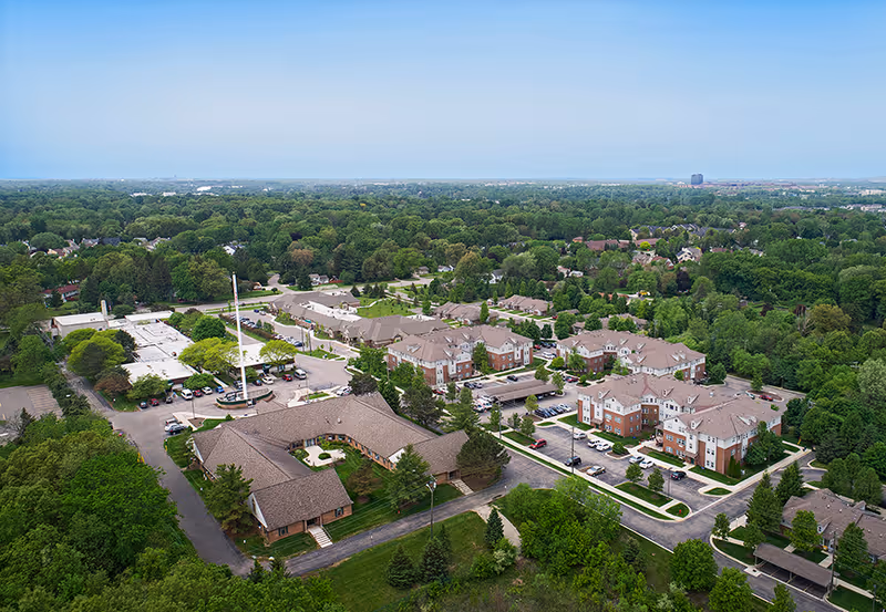 Aerial view of the American House Village of Rochester senior living facility surrounded by trees and greenery. The image shows multiple residential buildings with parking lots, roads, and landscaped areas under a clear blue sky.