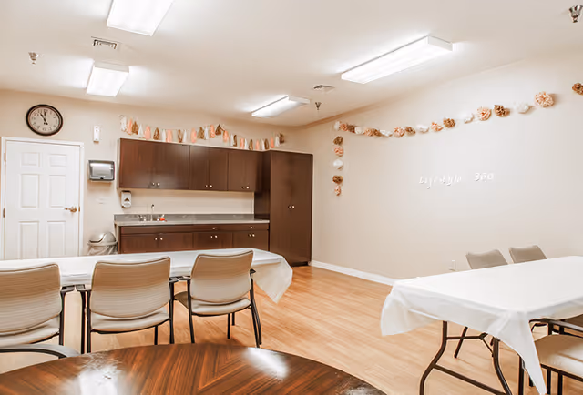 A bright room with wooden flooring featuring two tables covered with white tablecloths and several chairs around them. The back wall has dark wooden cabinets with a countertop and a sink. Above the cabinets, there is a decorative garland hanging along the walls. A clock is mounted on the wall above a closed white door. The words 'Lifestyle 360' are displayed on the right wall.