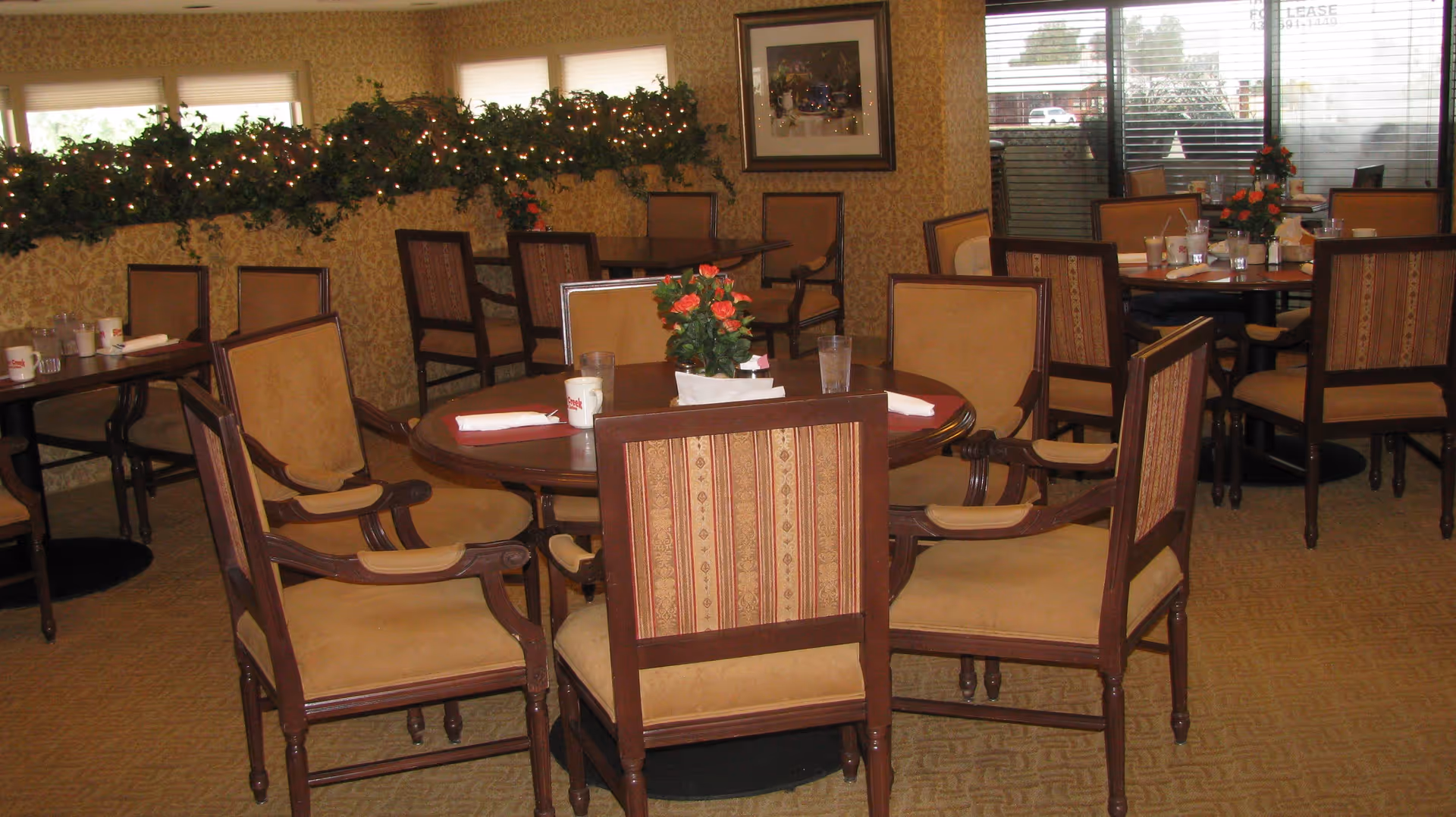 Dining room with round and rectangular wooden tables surrounded by cushioned chairs with patterned upholstery. Tables are set with napkins, glasses, mugs, and small flower arrangements. The room has patterned wallpaper, a decorative garland with lights on a ledge, and large windows with blinds letting in natural light.