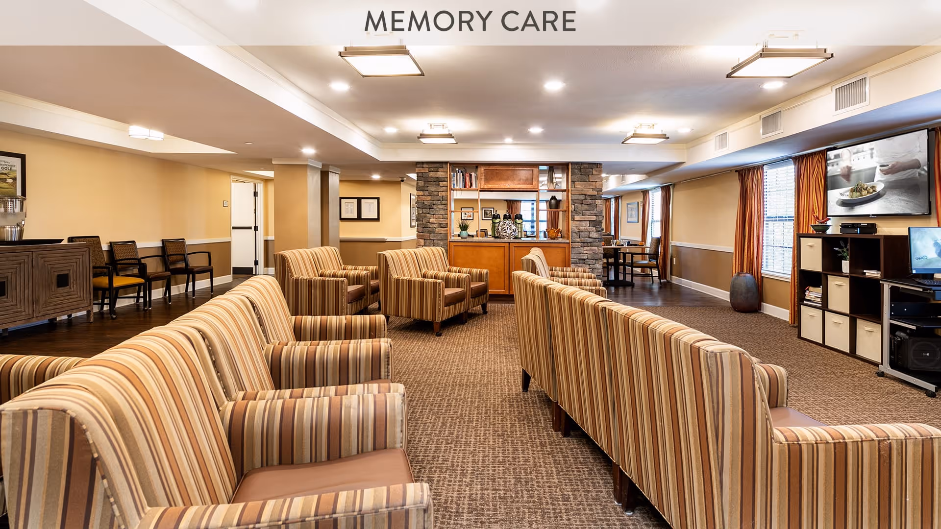 A spacious memory care common room with striped upholstered armchairs and sofas arranged in rows facing a television mounted on the wall. The room features warm beige walls, carpeted flooring, a wooden cabinet with shelves, and windows with orange curtains allowing natural light.