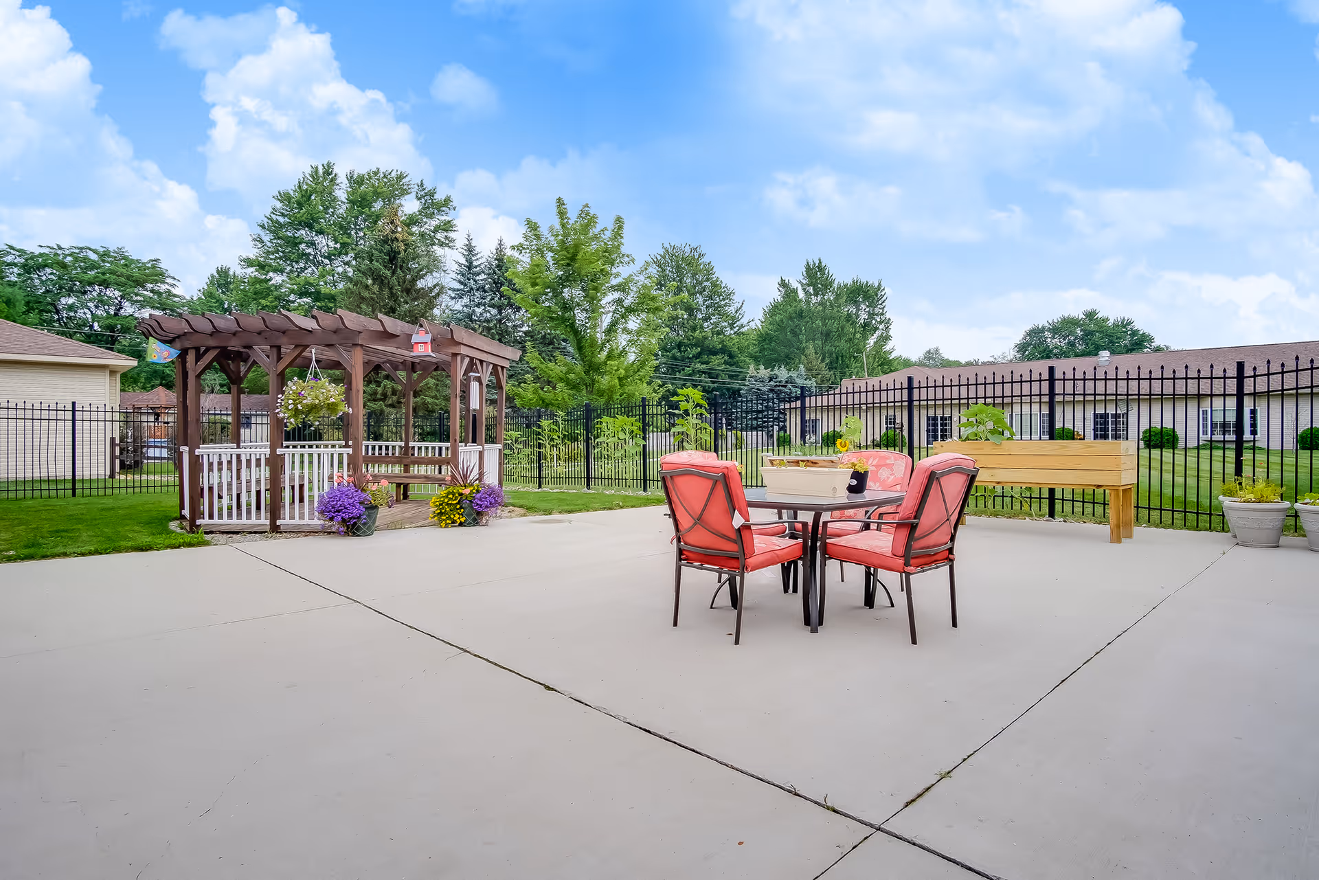 Outdoor patio with a table and red-cushioned chairs, a wooden pergola, planters, and a fenced lawn with buildings in the background.