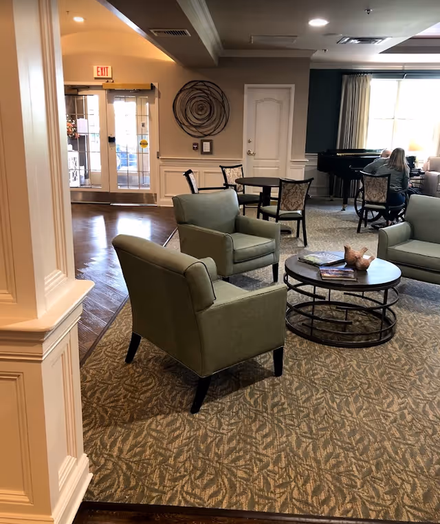 A cozy living room area in Avalon Assisted Living at Hillsborough featuring green upholstered armchairs arranged around a round coffee table with decorative items and books. In the background, there is a small dining table with chairs, a piano with two people seated nearby, and a glass door exit with an illuminated exit sign above it. The room has carpeted flooring with a patterned design and neutral-colored walls with decorative molding.