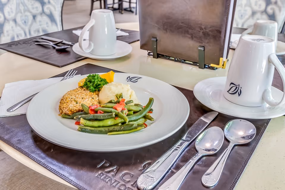 A dining table set with a plate of food including green beans with red peppers, mashed potatoes, a breaded patty, and a garnish of parsley. The table also has a white coffee cup on a saucer, silverware including a fork, knife, and two spoons, and a menu holder in the background. The placemat has the text 'PACIFIC SENIOR' partially visible.