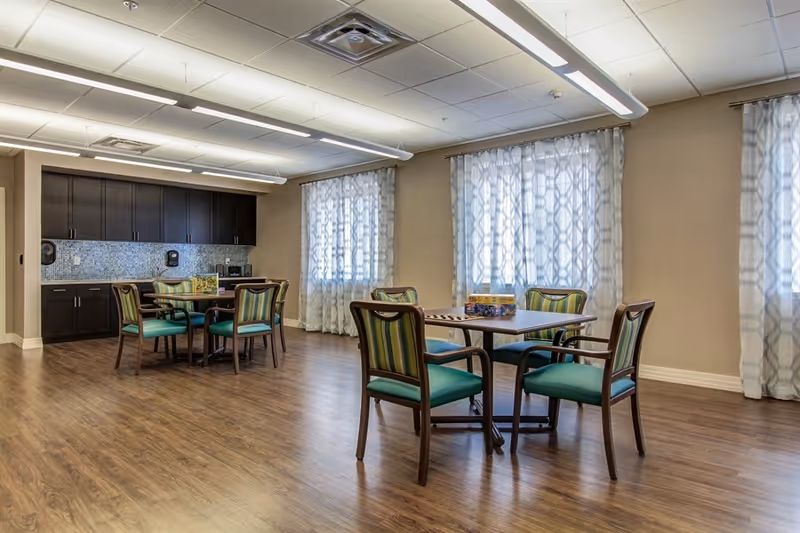 A spacious room with wooden flooring featuring two square tables each surrounded by four chairs with green and striped upholstery. The room has large windows with patterned sheer curtains allowing natural light to enter. Along one wall, there are dark wood cabinets with a mosaic tile backsplash and countertop, equipped with a microwave and other small appliances. The ceiling has modern fluorescent lighting fixtures.