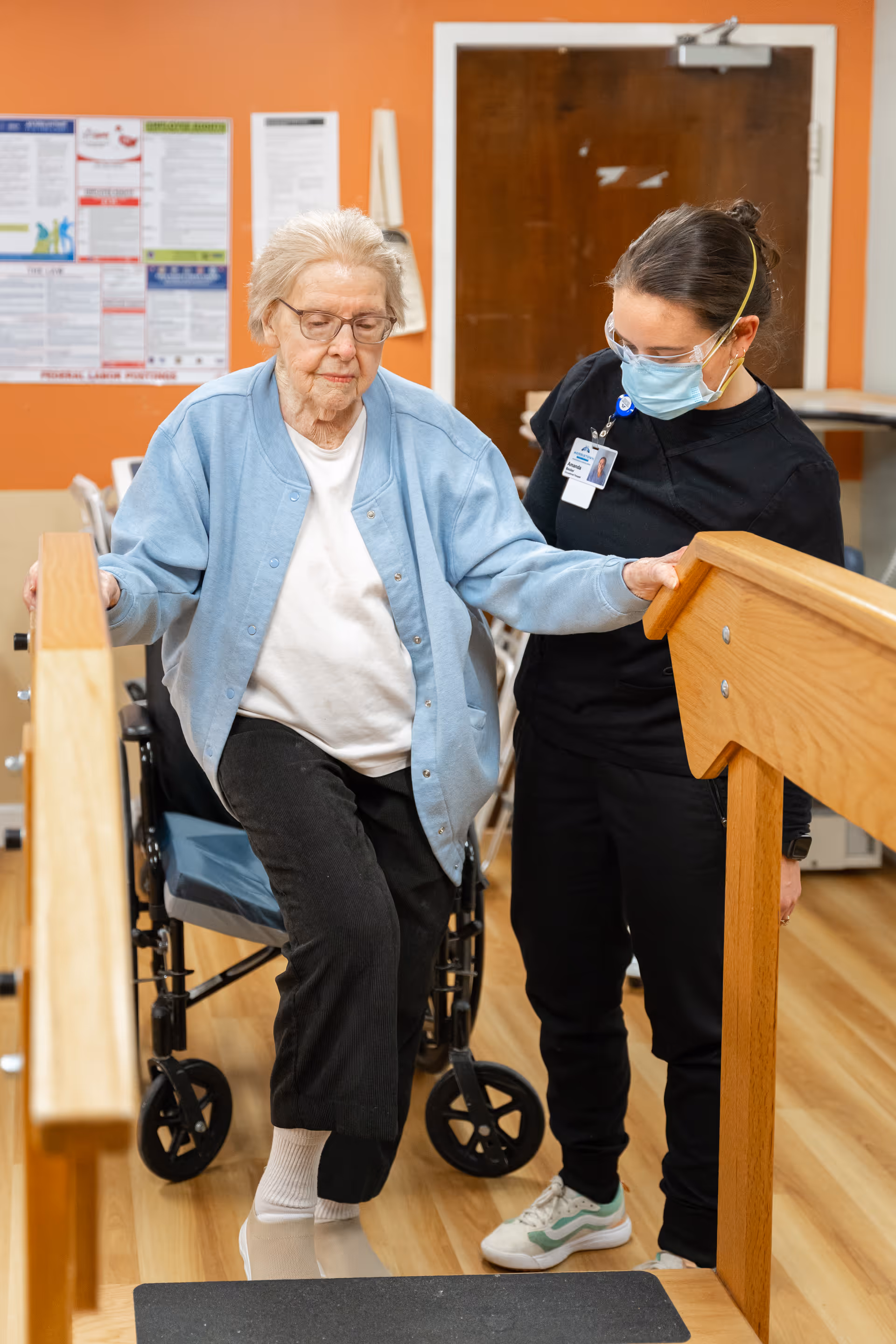 An elderly woman wearing glasses and a light blue cardigan is practicing walking exercises with the assistance of a healthcare worker wearing a mask and black scrubs. The elderly woman is holding onto wooden handrails and lifting one leg while a wheelchair is positioned behind her. The setting appears to be an indoor rehabilitation or nursing facility with an orange wall and informational posters in the background.