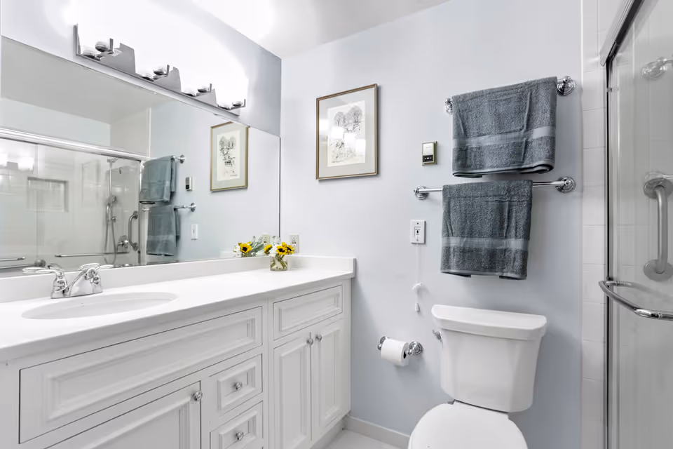 A clean, modern bathroom with a white vanity cabinet, a large mirror above the sink, two gray towels hanging on a double towel rack, a toilet, and a glass-enclosed shower with a grab bar. There is a framed picture on the wall and a small vase with yellow flowers on the vanity.