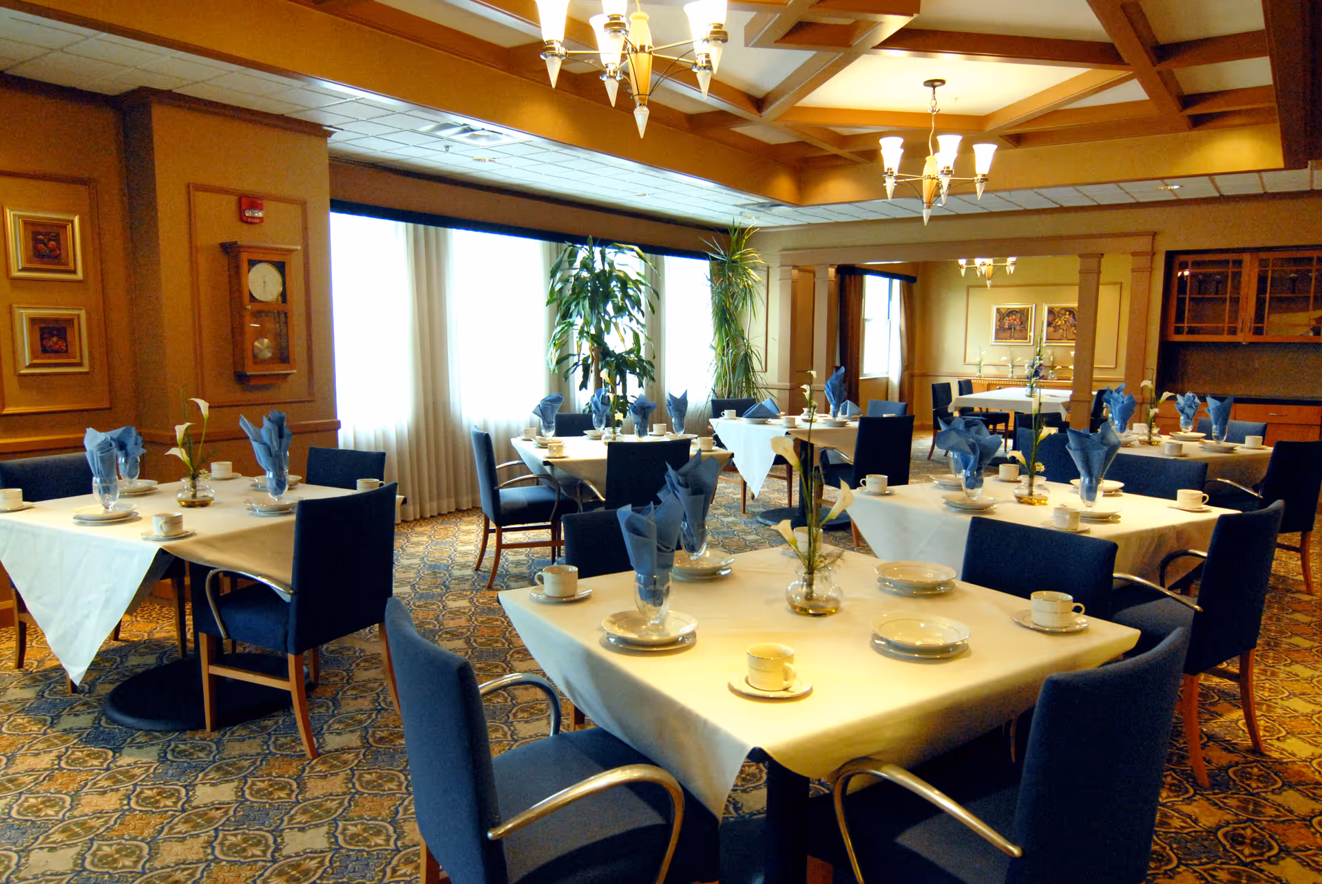 A well-lit dining room with multiple tables covered in white tablecloths, each set with blue napkins, white plates, cups, and small vases with flowers. The room has blue upholstered chairs, patterned carpet, large windows with curtains, and warm lighting from chandeliers on the ceiling.