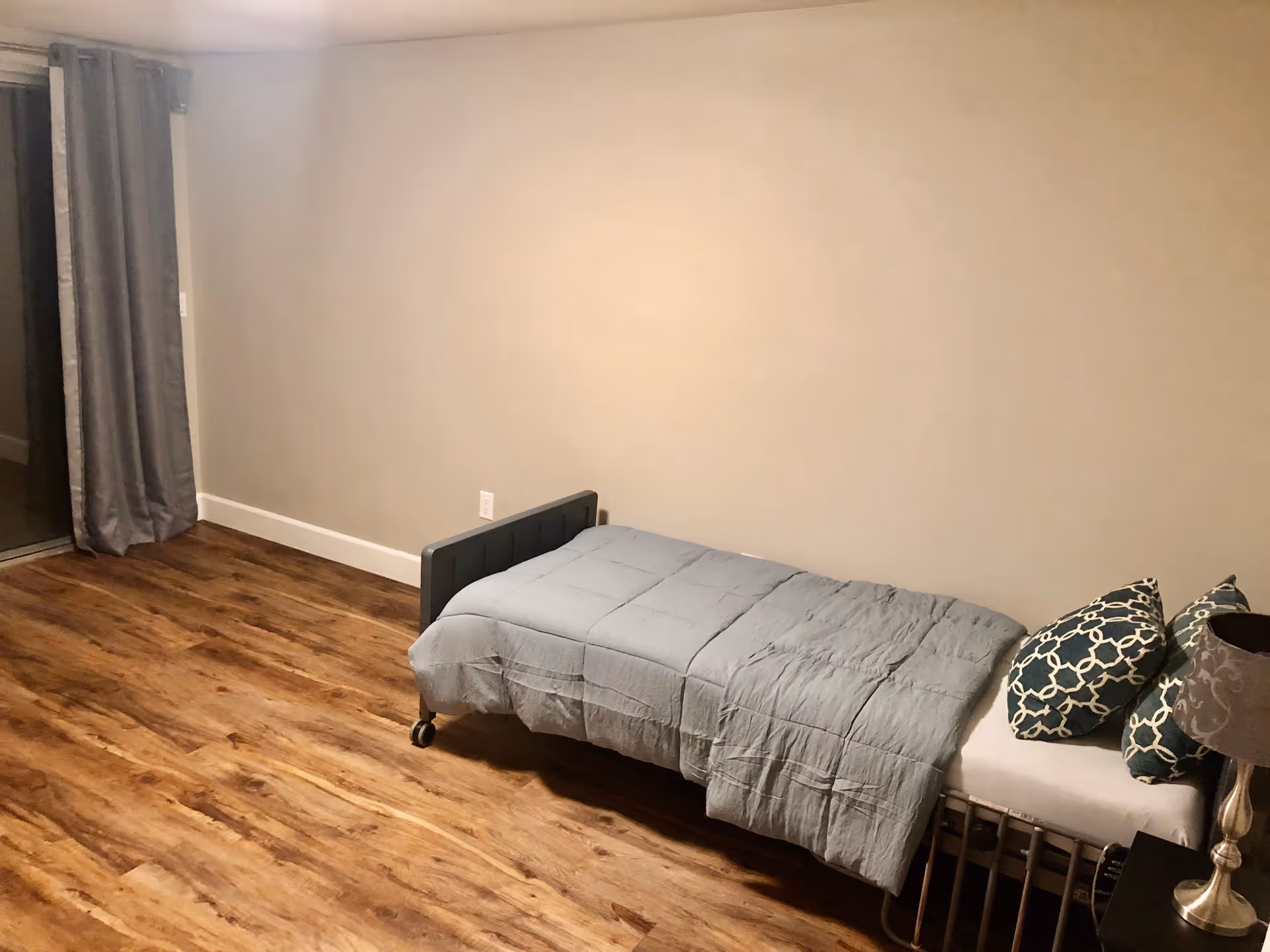 A simple bedroom with a single bed covered with a gray comforter and two patterned pillows. The room has wooden flooring, beige walls, a small bedside table with a lamp, and gray curtains covering a sliding glass door.