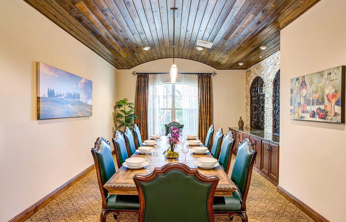 A formal dining room with a long wooden table set for twelve people with white plates and wine glasses. The chairs have green upholstery and wooden frames. The room features a wooden plank ceiling, a large window with sheer and patterned curtains, two paintings on the walls, a potted plant, and a sideboard with decorative items.
