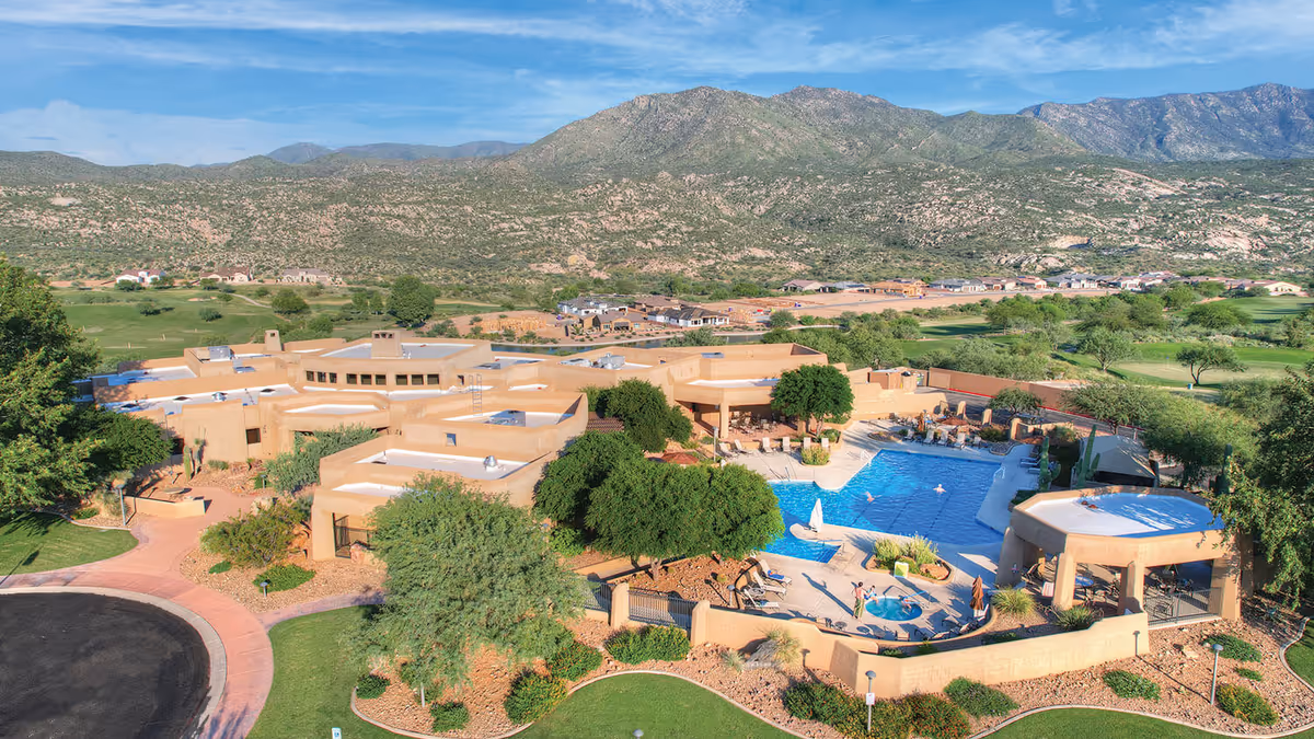 Aerial view of a Southwestern-style senior living complex with tan buildings, a central swimming pool and patios, and mountains in the background.