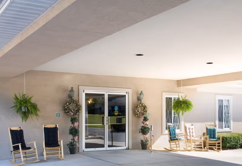 Covered entrance area of a senior living facility with glass double doors, two hanging green plants, floral wreaths on either side of the doors, and four wooden rocking chairs with cushions arranged on the concrete floor.