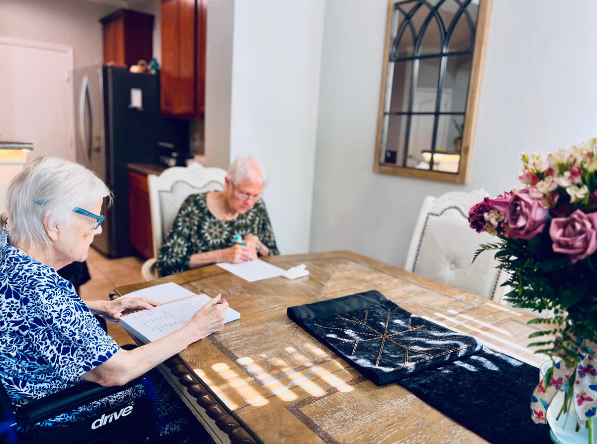 Two elderly women sitting at a wooden dining table in a well-lit room. One woman is in a wheelchair wearing a blue patterned shirt and glasses, writing in a notebook. The other woman, wearing a green patterned top, is also writing on a piece of paper. The room has a kitchen area with wooden cabinets and a refrigerator in the background. A decorative mirror hangs on the wall, and a vase with purple and pink flowers is on the table.