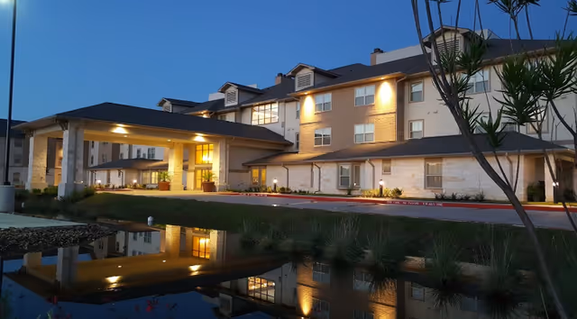 Front entrance of a three-story retirement facility at dusk with an illuminated porte-cochère and its reflection in a pond.
