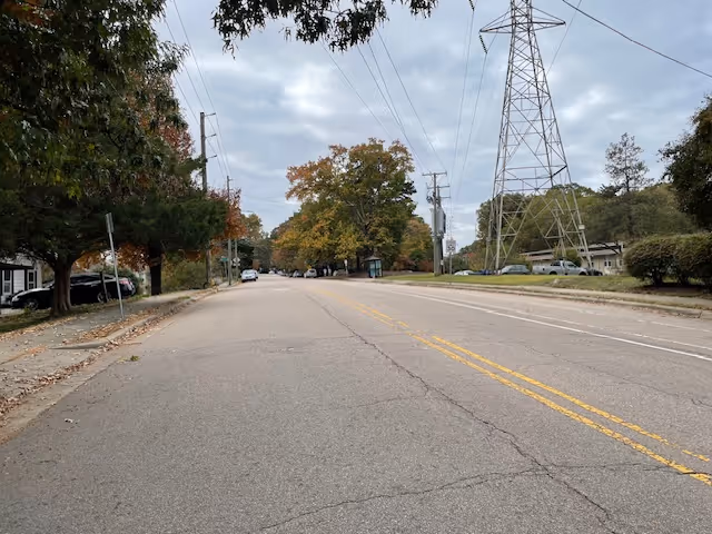 A wide street lined with trees on both sides, some showing autumn colors. There are cars parked along the left side of the street and a large electrical transmission tower on the right side. The sky is cloudy and overcast.