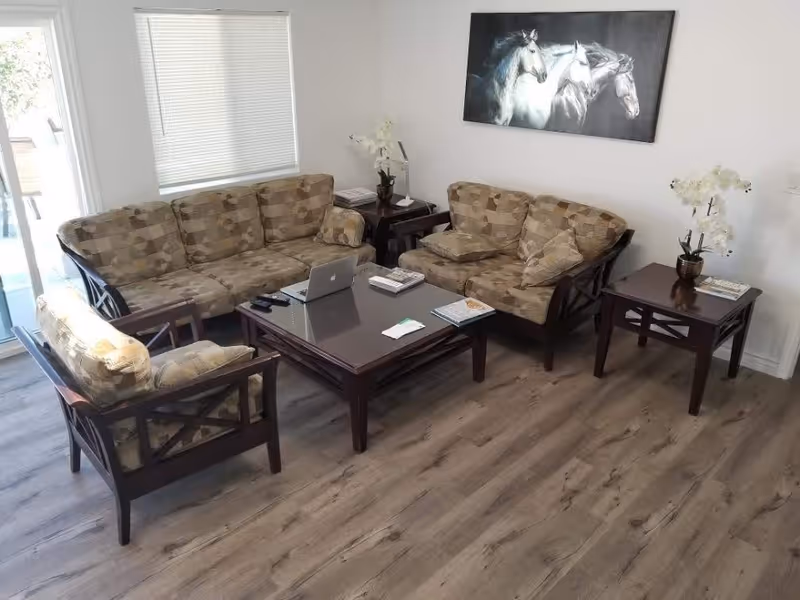 A living room with a three-seater sofa, a two-seater sofa, and a single armchair, all with patterned cushions. There is a dark wooden coffee table in the center with a laptop, remote controls, and some books on it. Two matching side tables each hold a white orchid plant and some books. A painting of three white horses hangs on the wall above the two-seater sofa. The room has wood flooring and a window with closed blinds.