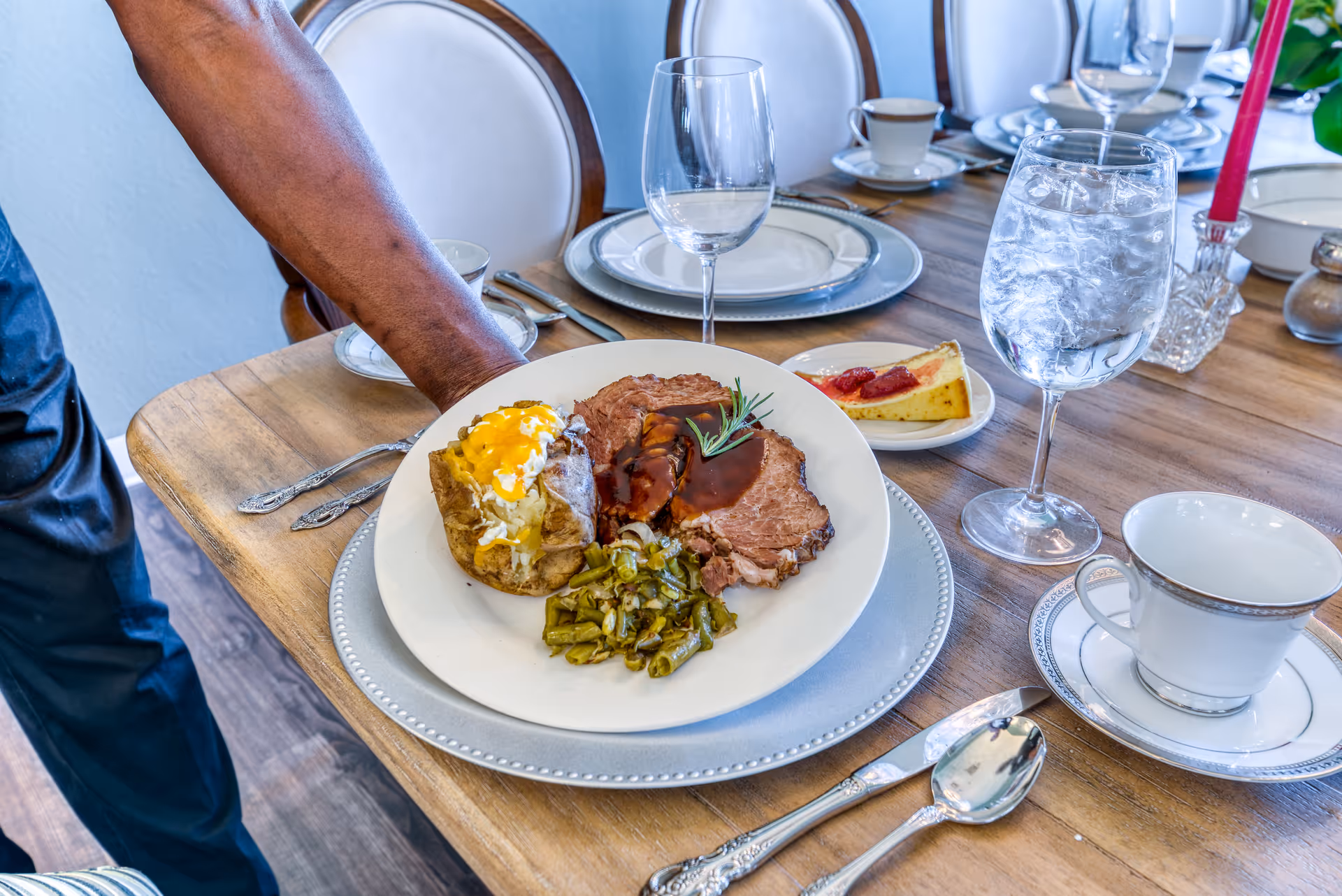 A hand placing a plate of food on a dining table set with plates, silverware, a glass of water, a wine glass, and a cup. The plate contains a baked potato with cheese, slices of roast beef with gravy, and green beans. A slice of cheesecake with strawberry topping is on a small plate in the background.