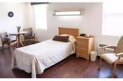 Bright bedroom with a single bed, nightstand, two chairs and a small table on hardwood floors.