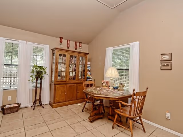 Small dining area with a round wooden table, chairs, a china cabinet, and windows with white curtains.