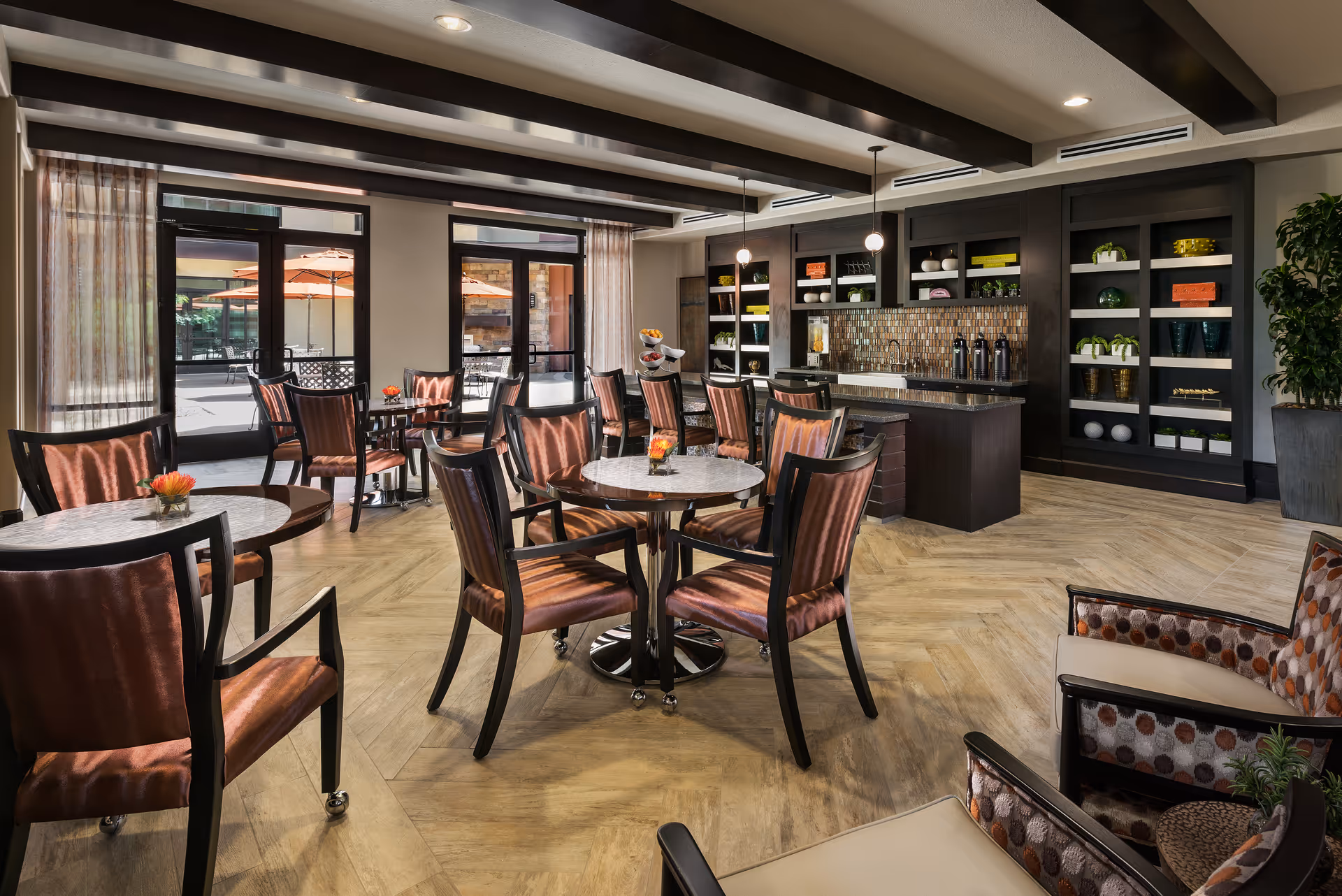 A well-lit dining area with multiple round tables and cushioned chairs arranged neatly. The room features large windows and glass doors leading to an outdoor patio with umbrellas. There is a counter with a backsplash and shelves holding decorative items and plants. The floor has a wood-like pattern, and the ceiling has exposed beams with hanging pendant lights.