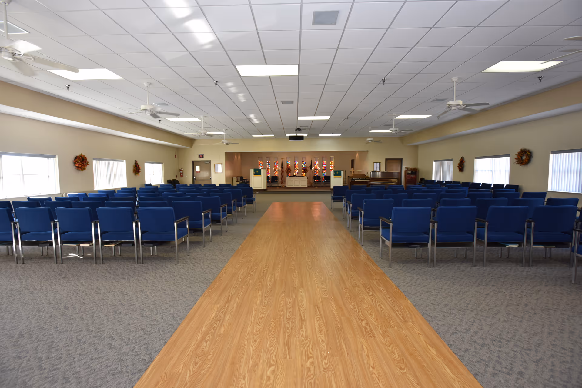 A large room with rows of blue chairs arranged on either side of a central wooden aisle. The room has a high ceiling with ceiling fans and fluorescent lights. At the far end, there is a stage area with stained glass windows and a piano to the right. The walls have windows with blinds and decorative wreaths.